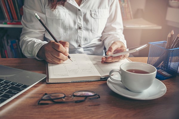 person sitting at desk writing in notebook and looking at phone