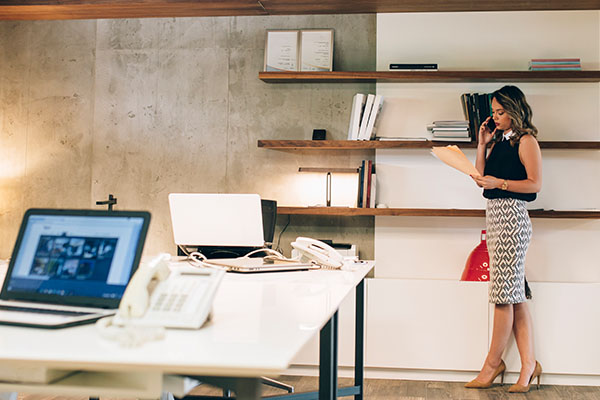 woman standing by desk looking at paperwork on a phone call